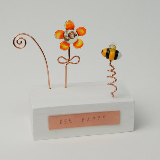 White wooden block with orange glass flower and bee with copper plaque reading Bee Happy